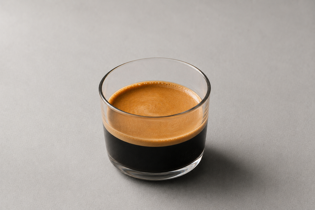 A clear glass tumbler filled with espresso, showing a rich golden-brown crema on top and dark coffee below, set against a solid gray background.