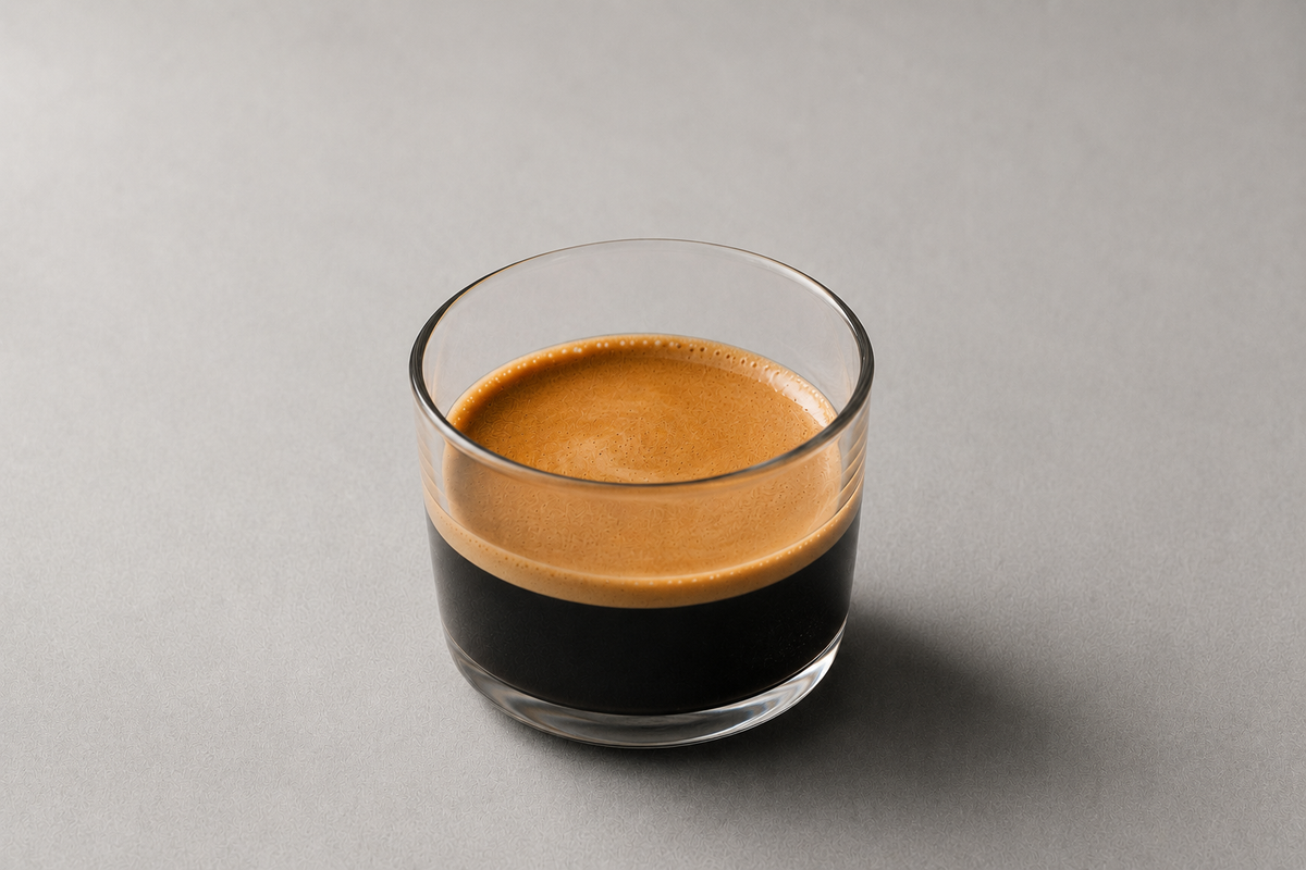 A clear glass tumbler filled with espresso, showing a rich golden-brown crema on top and dark coffee below, set against a solid gray background.