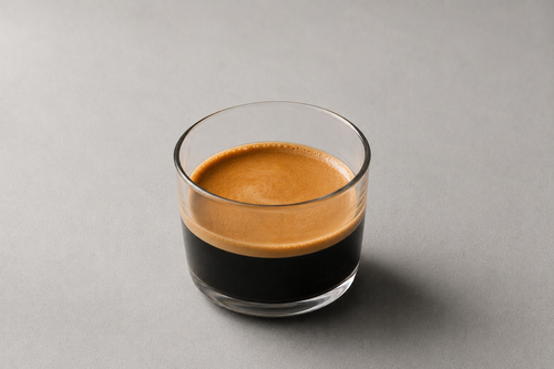 A clear glass tumbler filled with espresso, showing a rich golden-brown crema on top and dark coffee below, set against a solid gray background.