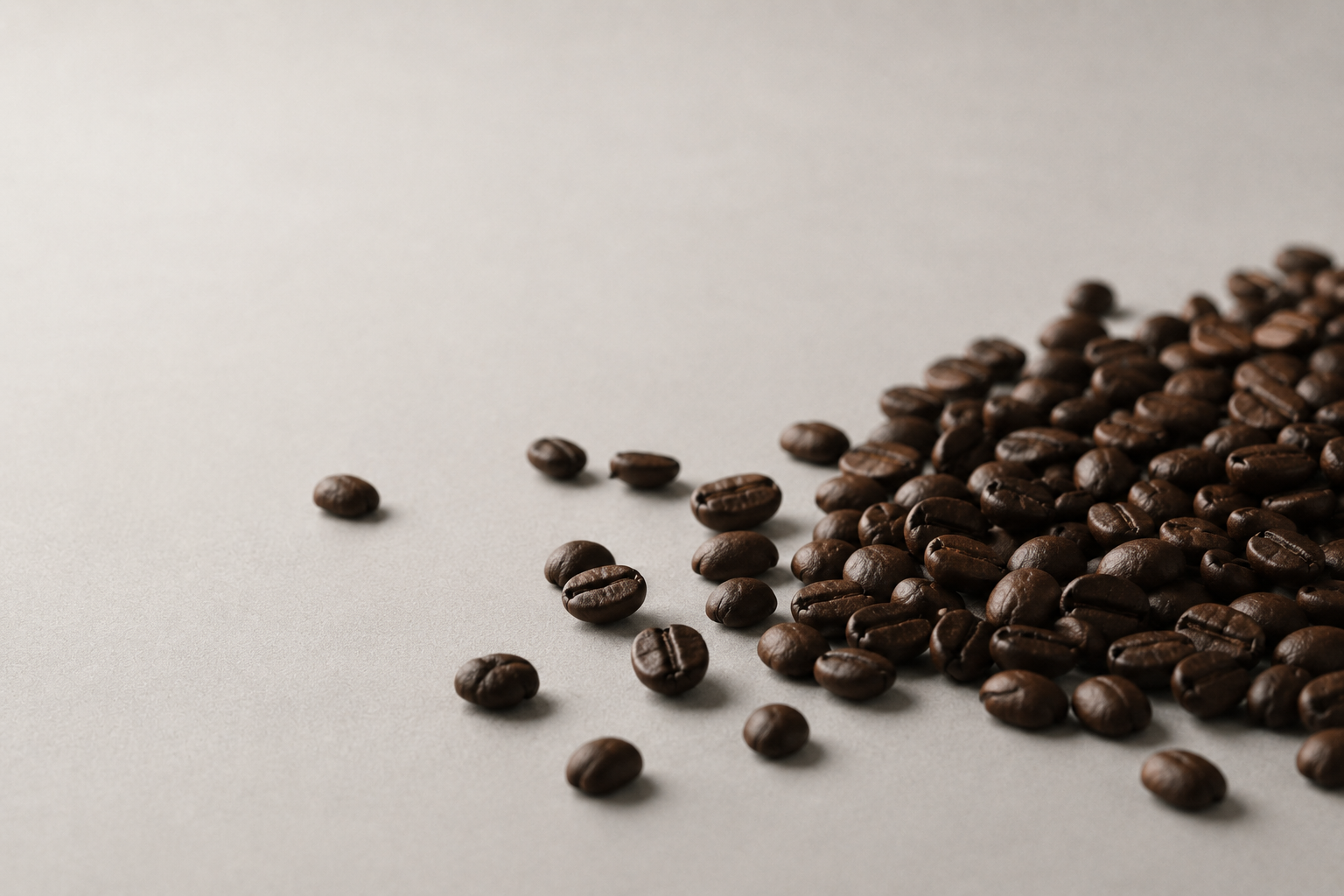 Dark roasted coffee beans scattered on a light gray surface, with a cluster concentrated on the right side and a few beans spaced out on the left.