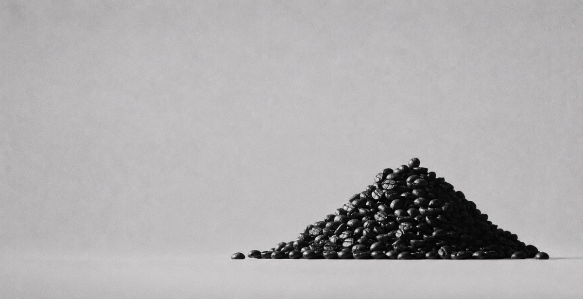 A pile of dark roasted coffee beans sits on a light gray surface against a matching light gray background.